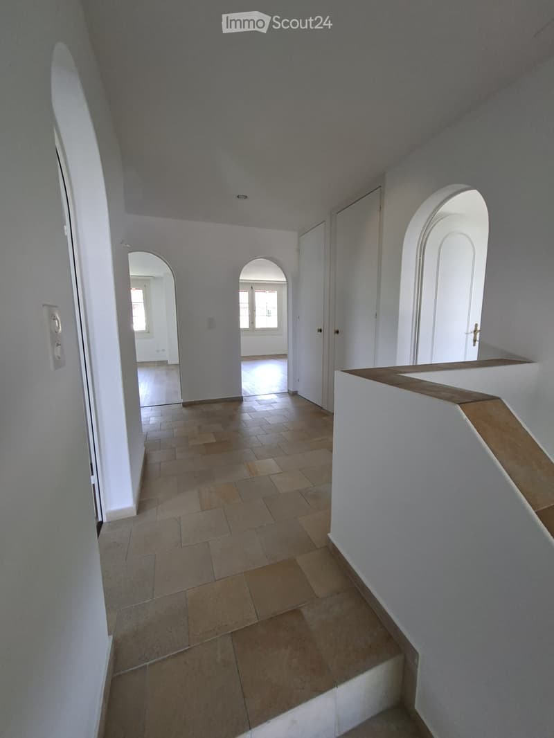 Empty hallway with tiled floor, white walls, arched doorways, and a wooden railing on the right side.