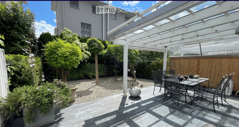 A well-maintained garden area with a covered patio, featuring a dining table and chairs, surrounded by greenery and trees.