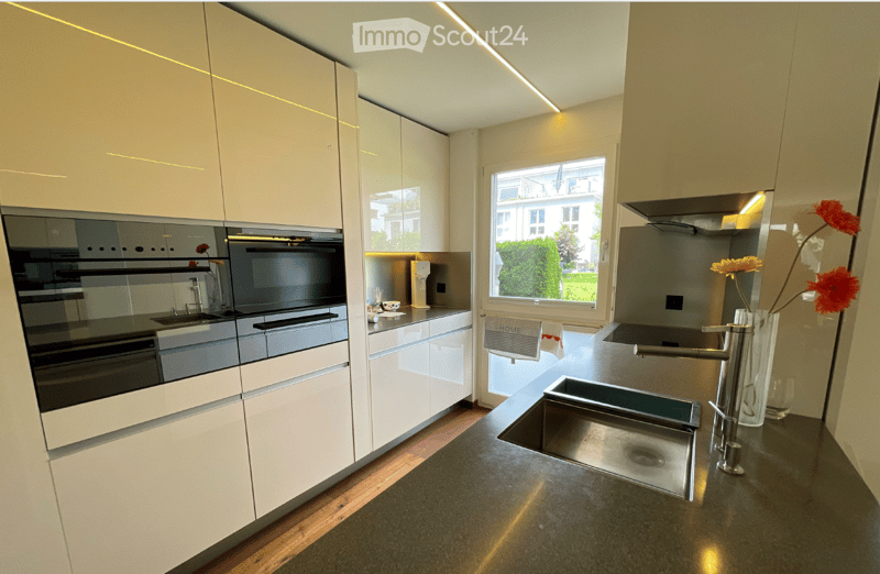 Modern kitchen with white cabinets, stainless steel sink, built-in oven, and a large window offering a view of the garden.