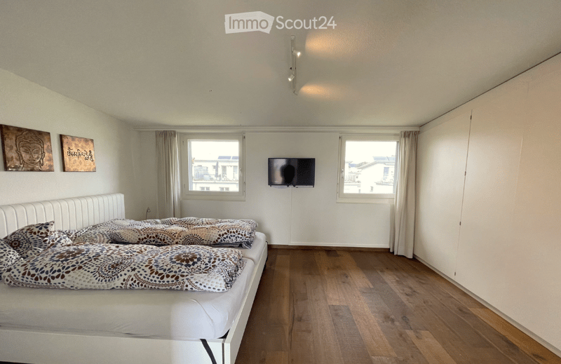 Modern bedroom with a white bed, patterned pillows and blanket, mounted TV, large windows with curtains, and artwork on the walls.