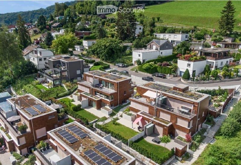 Aerial view of a residential area with modern houses, solar panels on rooftops, well-maintained gardens, and a scenic green landscape in the background.