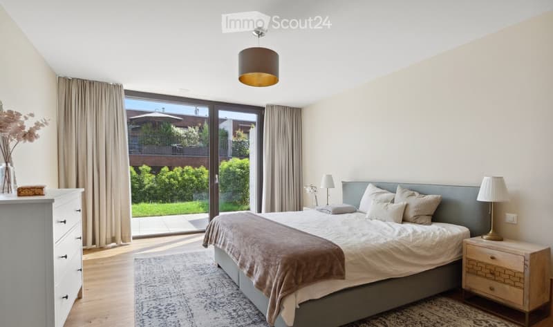 Modern bedroom with a large bed, nightstands, lamps, dresser, and a view of a garden through large windows.