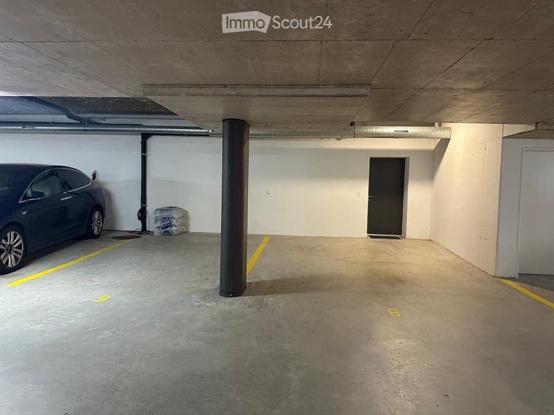 Underground parking area with one parked car, concrete floor, white walls, and a central support column.