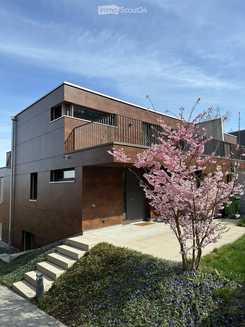 Modern two-story house with a brown facade, large windows, and a balcony on the upper level. A small tree with pink blossoms is in the front yard, along with neatly trimmed bushes and a small garden area.