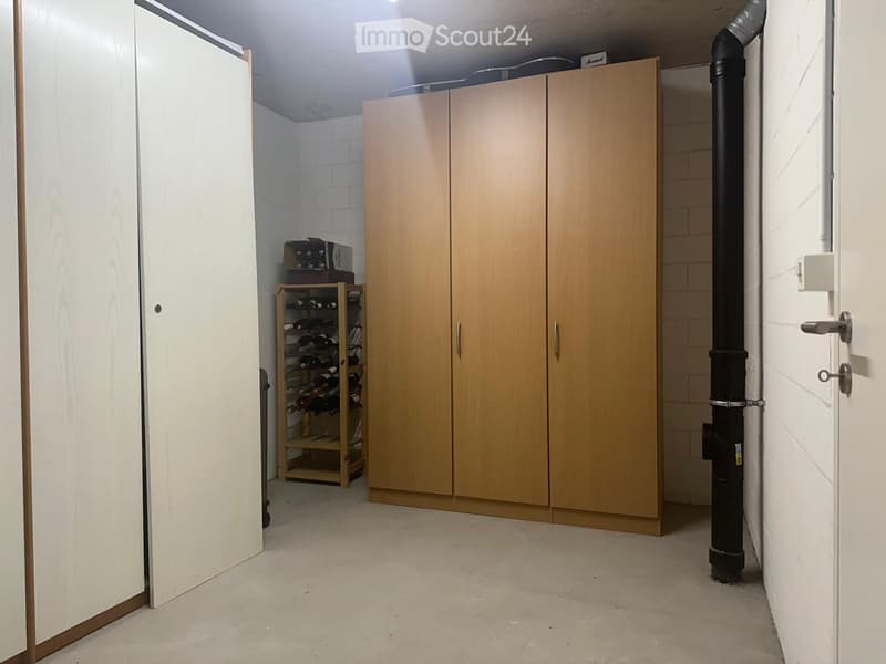 A small, organized storage room with a wine rack and a large wooden cabinet. The room has white brick walls and a concrete floor.