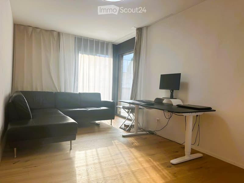 A modern living room with a black leather sofa, a standing desk with a laptop and screen, and large windows with sheer curtains.