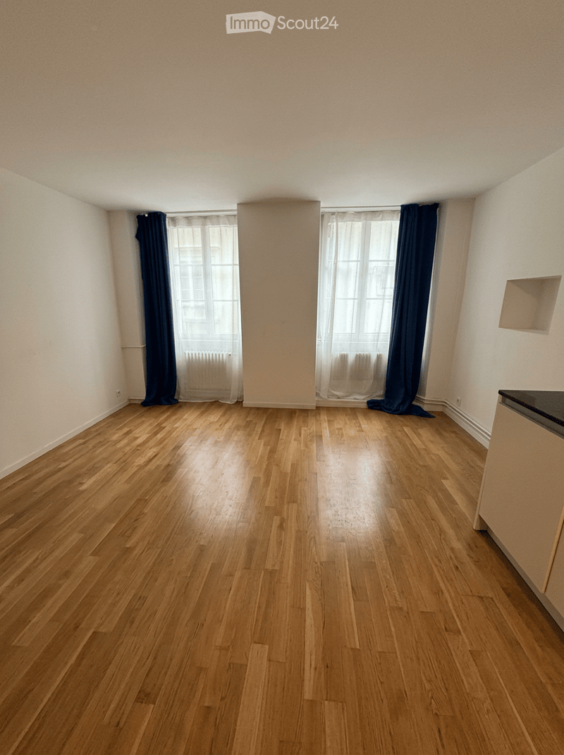 Empty room with wooden flooring, white walls, large windows with dark curtains, and a small built-in shelf on one wall.