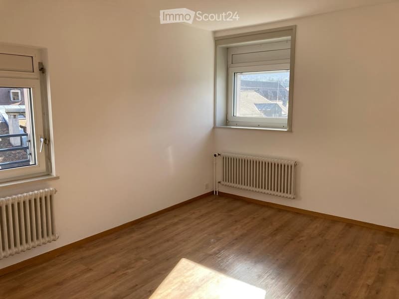 Empty room with wooden flooring, white walls, two windows, and radiators on both sides.