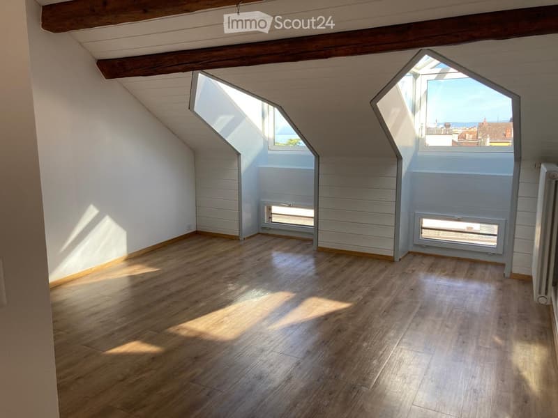 Empty attic room with wooden flooring, white walls, exposed wooden beams, and skylight windows offering a view of the sea and surrounding rooftops.