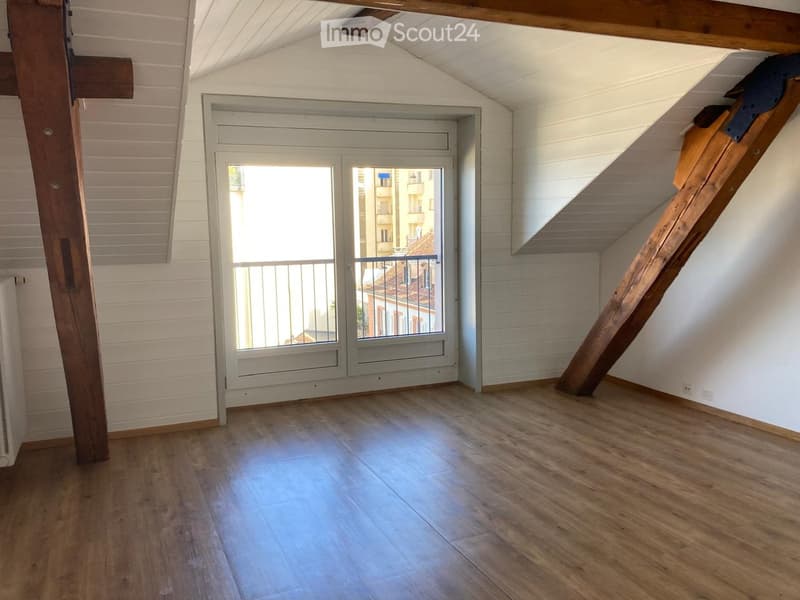 Empty attic room with wooden floors, white walls, exposed wooden beams, and a staircase leading up. Features a large window with a view of the city and a balcony door.