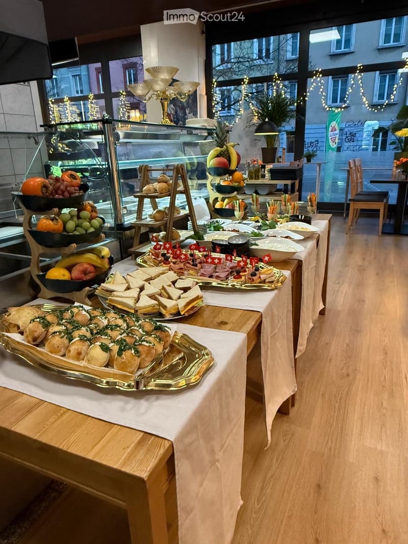 A buffet setup with various food items including sandwiches, fruits, and cakes arranged on tables covered with white tablecloths. The room has large windows with a view of the outside and is decorated with string lights.