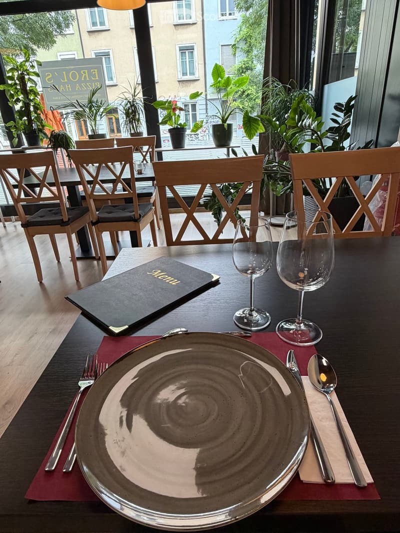 A restaurant setting with a table set for two, featuring a menu, wine glasses, a large plate, and silverware, surrounded by wooden chairs and potted plants.