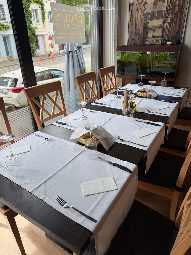 Restaurant interior with a long table set for dining, featuring wine glasses, cutlery, napkins, and bread. Wooden chairs surround the table, and large windows offer a view of the street outside.