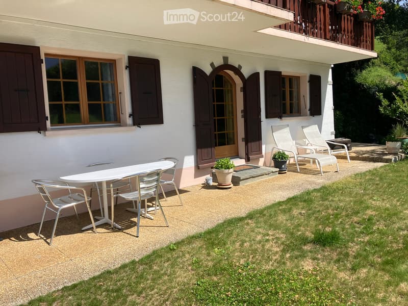 Outdoor patio area with a white table and multiple chairs, a small potted plant, and a grassy lawn. The patio is adjacent to a house with white walls, wooden shutters, and a wooden deck with additional seating.