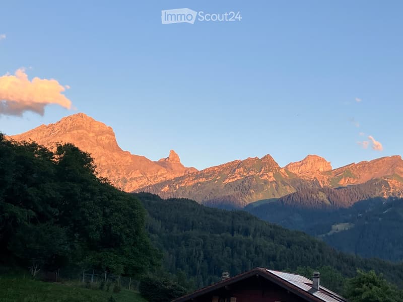 A traditional wooden house with a sloped roof and chimney, situated in a lush green valley surrounded by towering mountains under a clear blue sky.