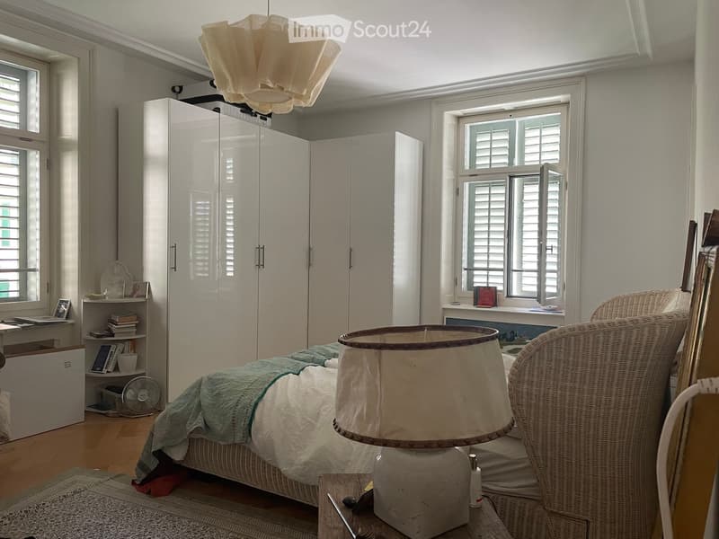 A well-lit bedroom with a large bed, a wicker chair, a wardrobe, a table lamp, a fan, a bookshelf, and a laptop on a desk.