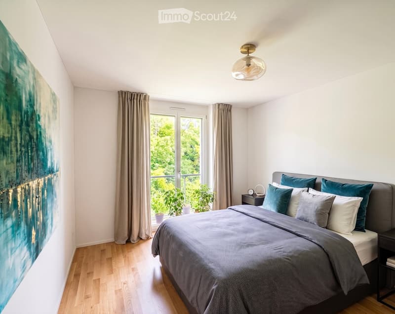 Modern bedroom with a large bed, grey bedding, blue pillows, nightstand, and large window with curtains. A painting adorns the wall, and the room has wooden flooring.