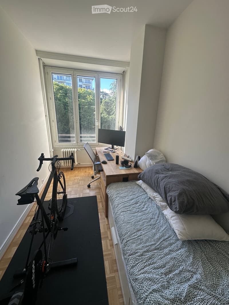 A small, furnished bedroom with a single bed, desk, chair, computer, and bicycle. The room has wooden flooring and large windows with a view of trees.