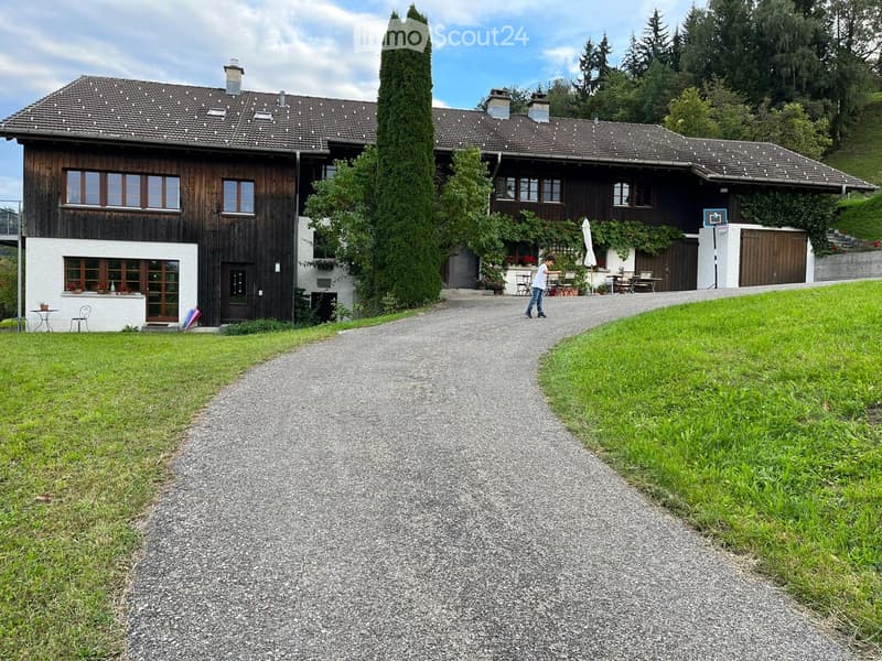 A two-story wooden house with a tiled roof, multiple windows, and a covered outdoor seating area. The house is surrounded by greenery and a gravel driveway.