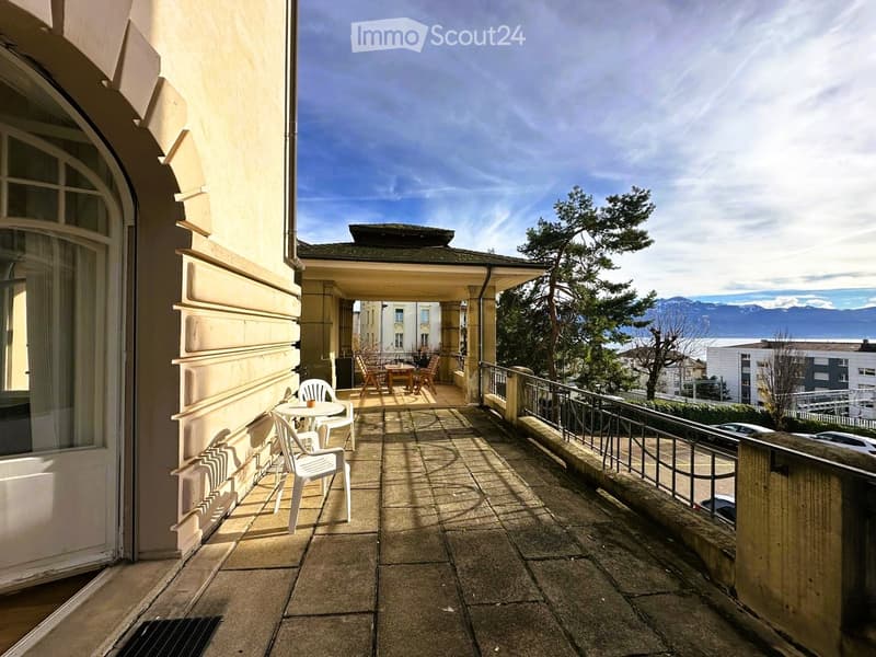 Balcony with tiled floor, white plastic chairs, small table, and a view of the city and mountains.