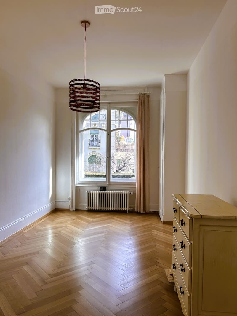 Empty room with wooden herringbone flooring, white walls, large arched windows with curtains, a radiator, and a decorative pendant light fixture.
