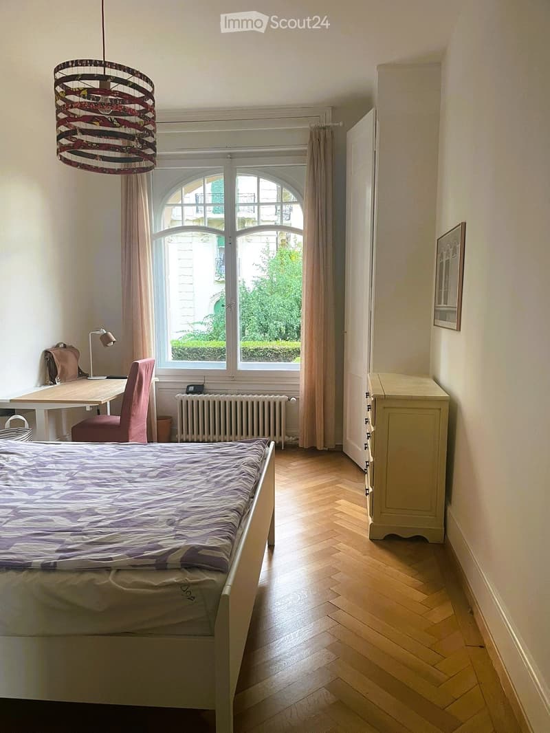 Cozy bedroom with wooden flooring, a single bed with a patterned duvet, a pink chair, a small desk with a lamp, a radiator, and a cabinet.