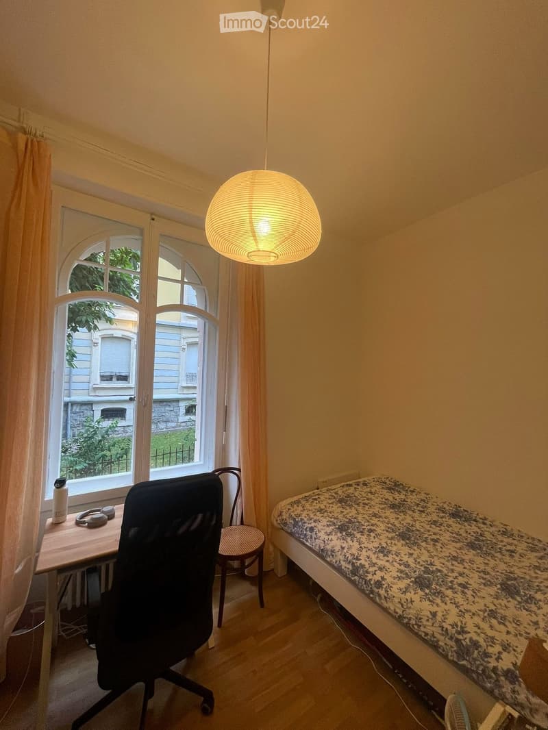 A small, cozy bedroom with a bed, a desk with a chair, a hanging lamp, and a window with curtains.