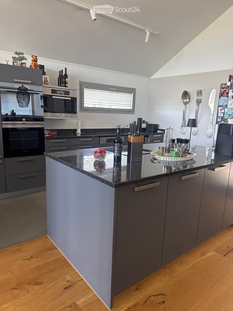 Modern kitchen with grey cabinets, granite countertop, stainless steel appliances, and various kitchen items including a bottle, bowl, and knife block.