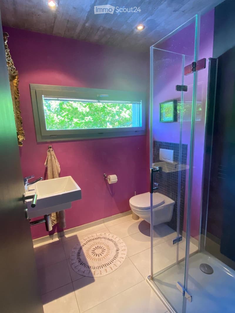 Modern bathroom with pink walls, white tiled floor, a sink, toilet, and a glass-enclosed shower. A large window provides natural light, and a towel hangs on the wall.