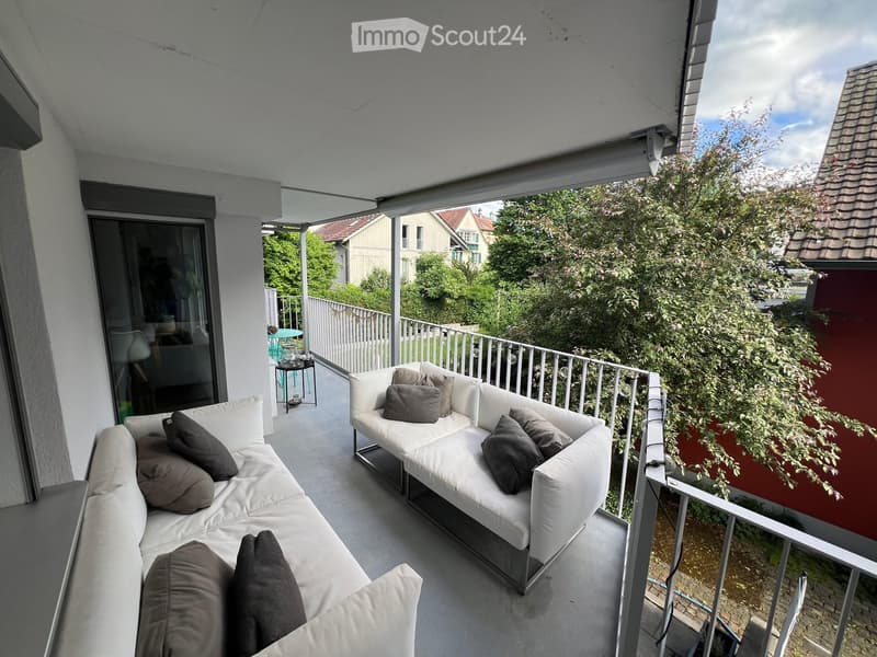 Balcony with white sofa and cushions, overlooking a green area with trees and neighboring houses.