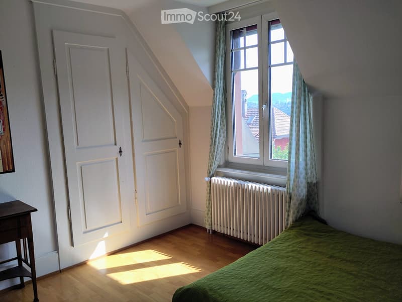 Bright attic bedroom with wooden flooring, a single bed with green bedding, a nightstand, and large windows with curtains offering a view of the roof and surrounding area.