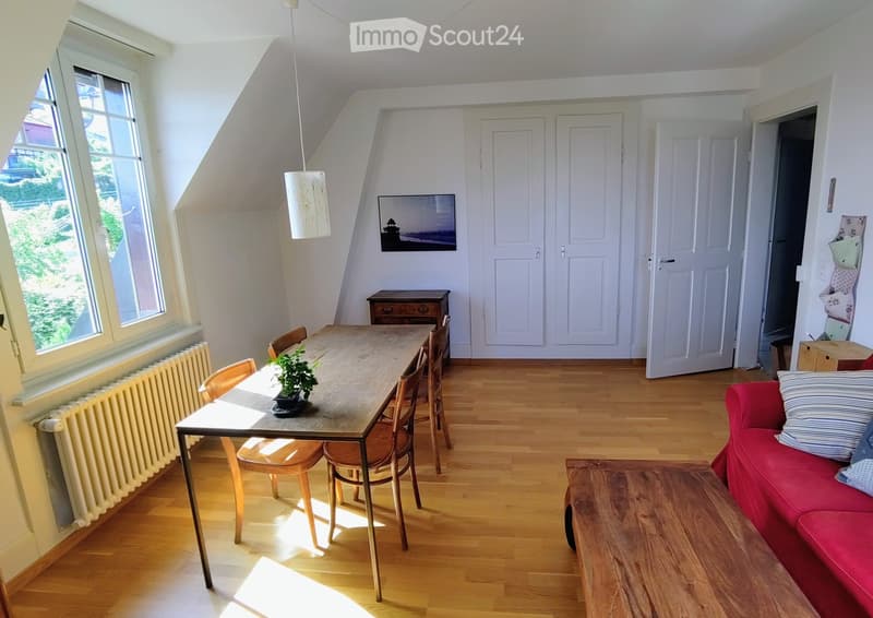 Bright living and dining area with wooden flooring, large windows, a red sofa, a wooden dining table with chairs, and a small potted plant.