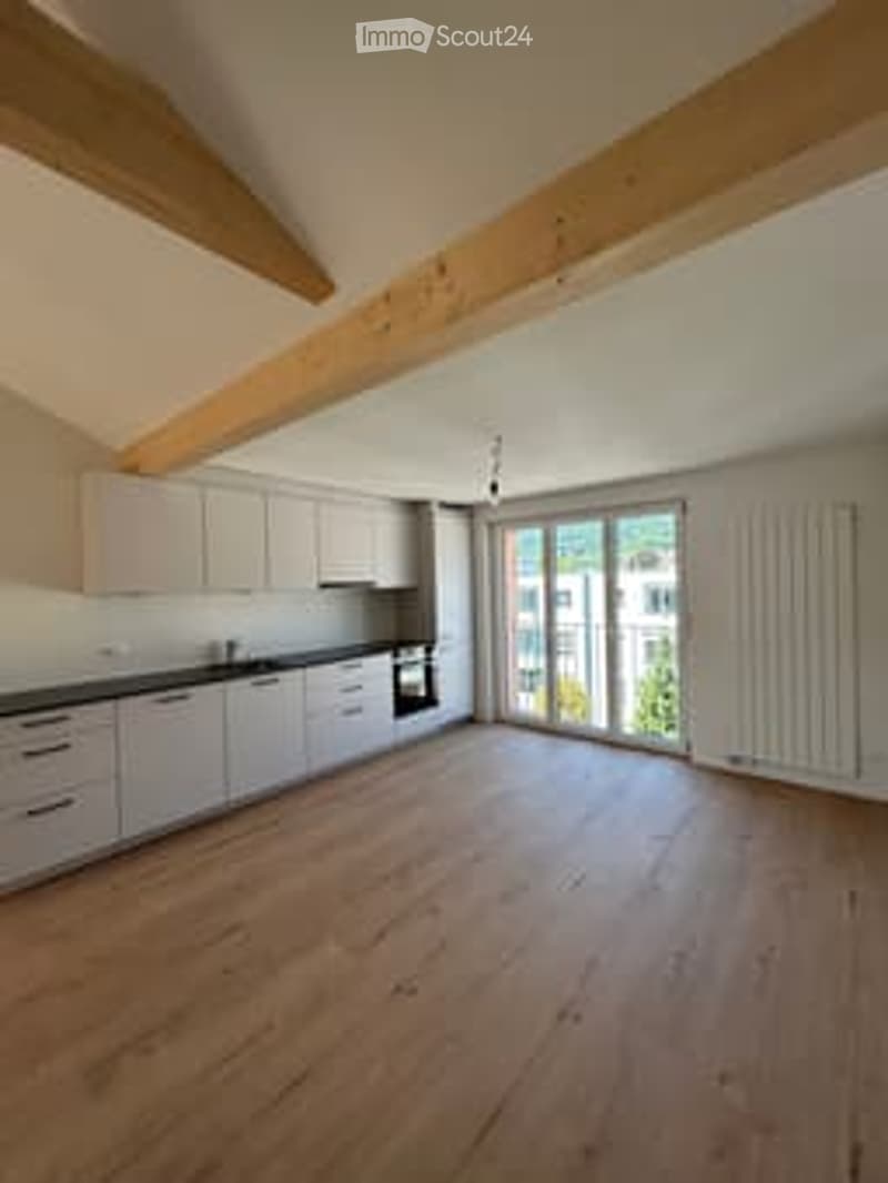 Bright, open kitchen with white cabinets, dark countertops, and large windows offering a view. The floor is made of light wood, and the space is completely empty with no furniture or appliances.
