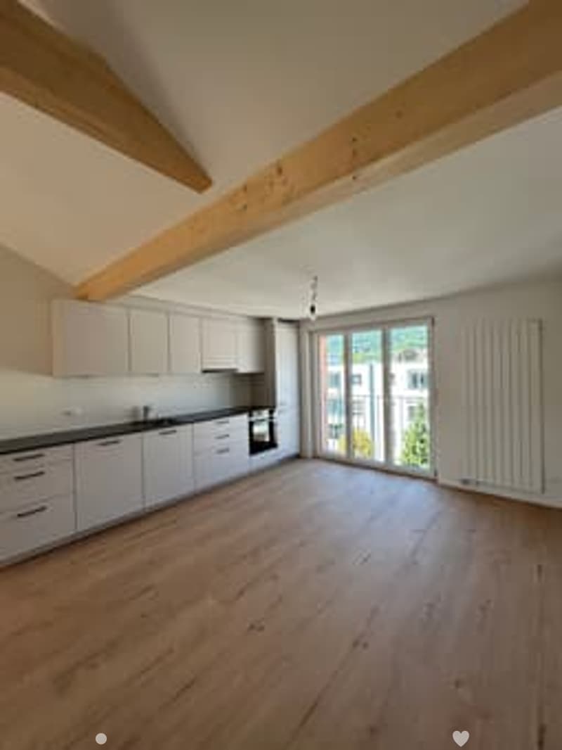 Bright, open kitchen with white cabinets, dark countertops, and large windows offering a view. The floor is made of light wood, and the space is completely empty with no furniture or appliances.