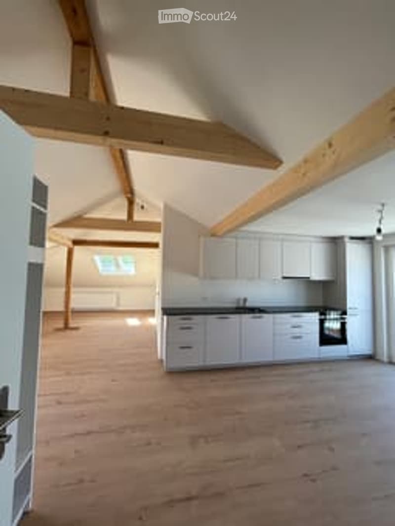 Bright, open kitchen with white cabinetry, stainless steel appliances, and exposed wooden beams on the ceiling. The floor is made of light wooden planks.