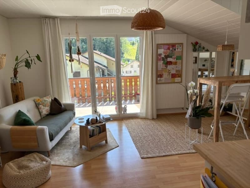 Bright living room with large windows and a balcony view, featuring a grey sofa, coffee table, ottoman, and hanging pendant lights.