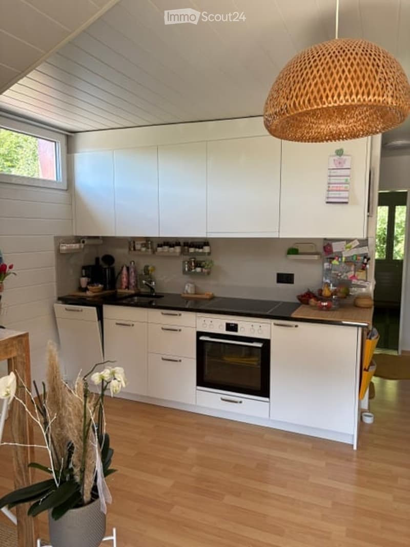 Modern kitchen with white cabinets, wooden flooring, and a central island. Features include a sink, oven, dishwasher, and a wicker pendant light hanging from the ceiling.
