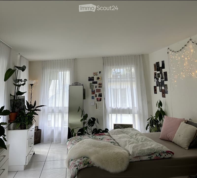 Cozy bedroom with a bed, floral duvet, fur throw, pink pillows, a mirror, a small dresser, and several potted plants.