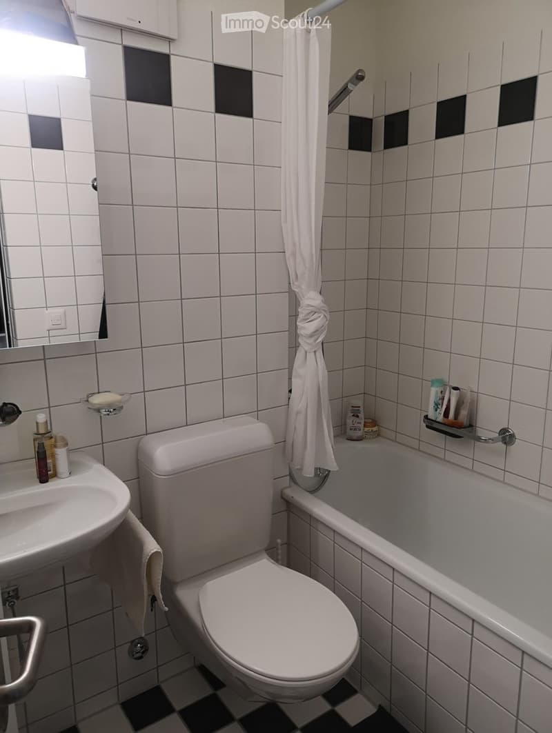 A small bathroom with white tiled walls, a toilet, a sink, a bathtub, and a mirror. A towel hangs on a rod, and various toiletries are placed on the sink and bathtub ledge.