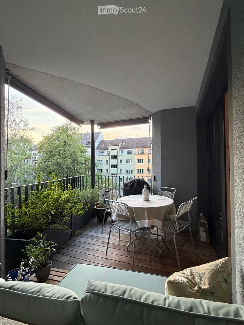 Balcony with wooden decking, round table with chairs, sofa with cushions, potted plants, and a covered barbecue.
