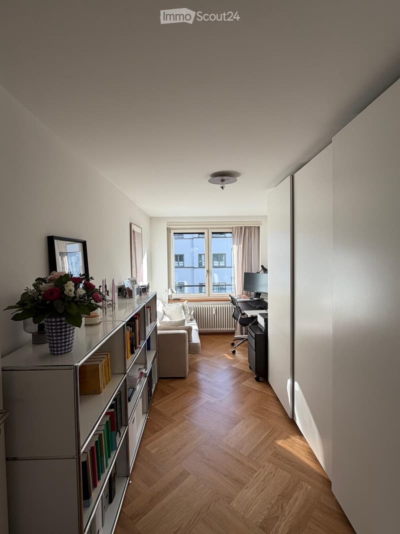 A well-lit room with wooden herringbone flooring, a sofa, bookshelf, desk with computer, and a flower vase on the shelf.