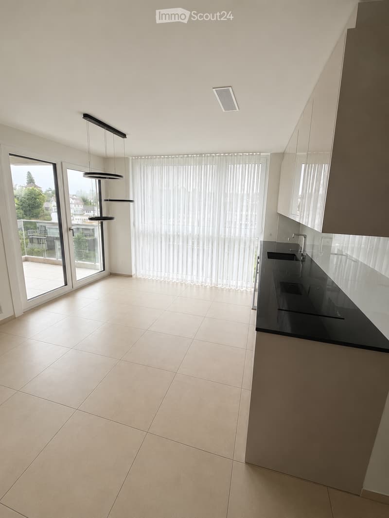Empty kitchen with tiled floor, white walls, black countertop, and large windows with sheer curtains. No furniture or appliances are present.