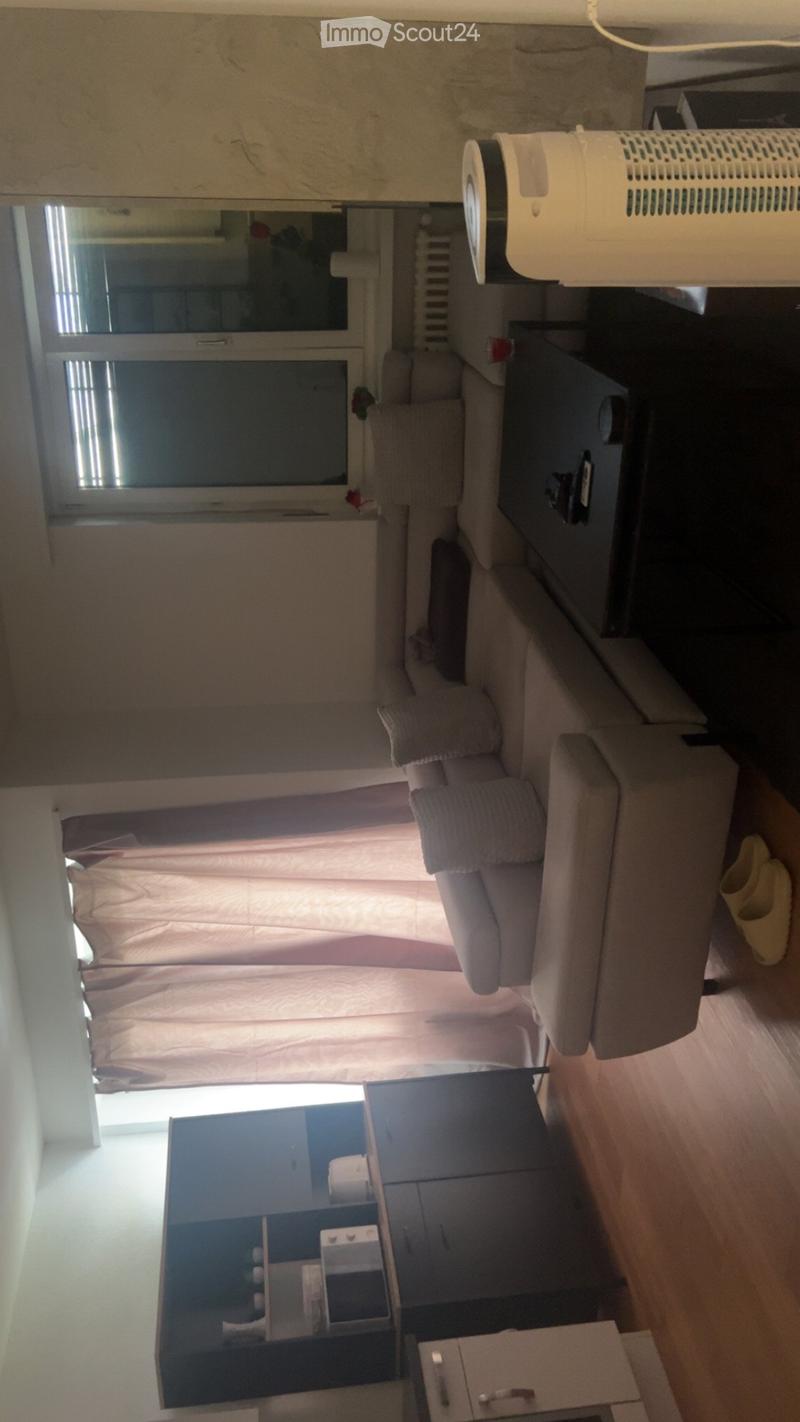 A cozy living room with a grey sofa, a small table, a chair, and a window with curtains. The room features a radiator and an air purifier on the wall.