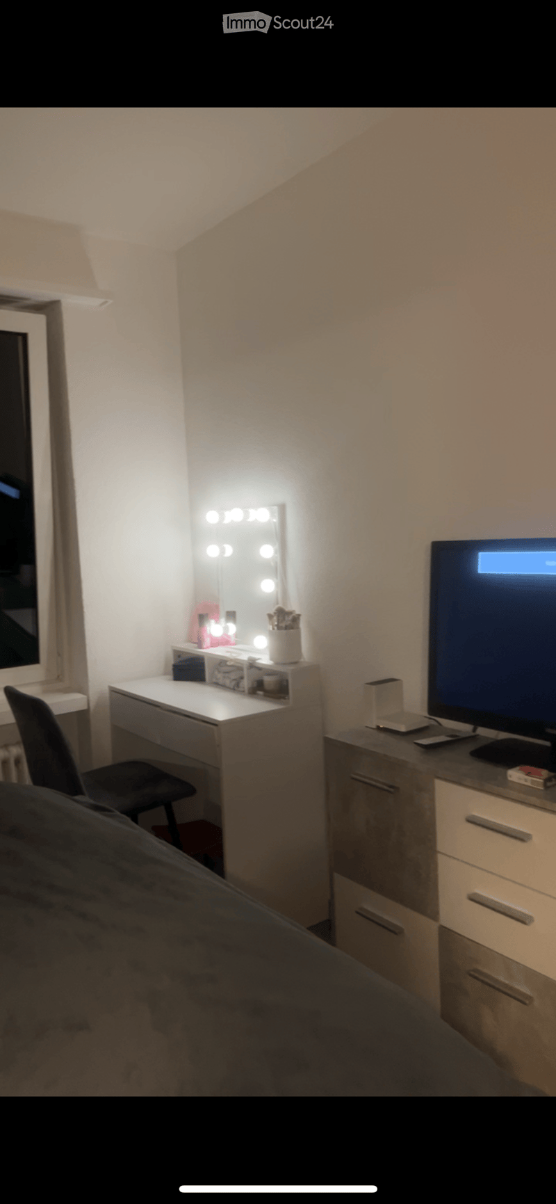 A cozy bedroom with a large mirror, vanity desk, chair, television, and drawer unit. The room features soft lighting and a comfortable bed.