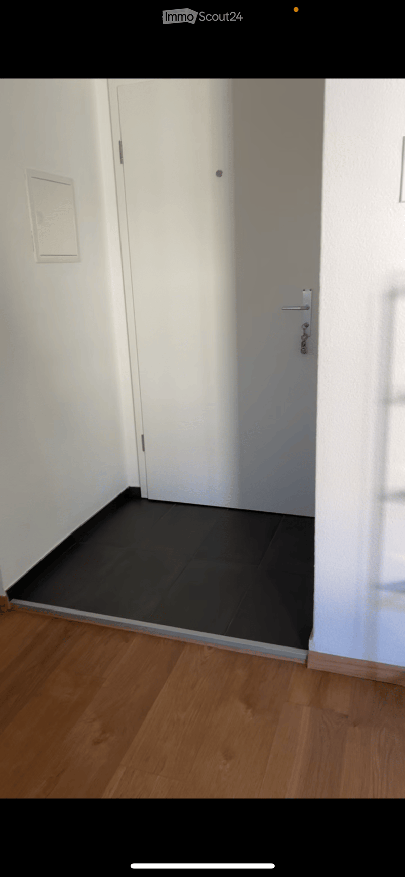 Empty hallway with wooden flooring and white walls, featuring a closed white door and a partially visible adjacent room with black tiled flooring.