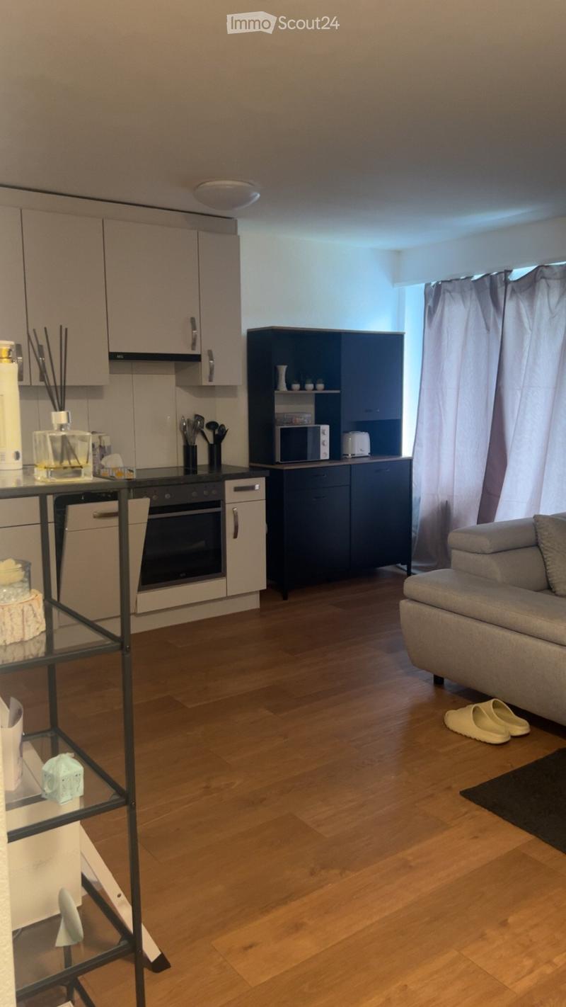A modern, open-plan living and kitchen area with light wooden flooring, a grey sofa, a glass table, and a kitchen area with an oven, microwave, and cabinets.