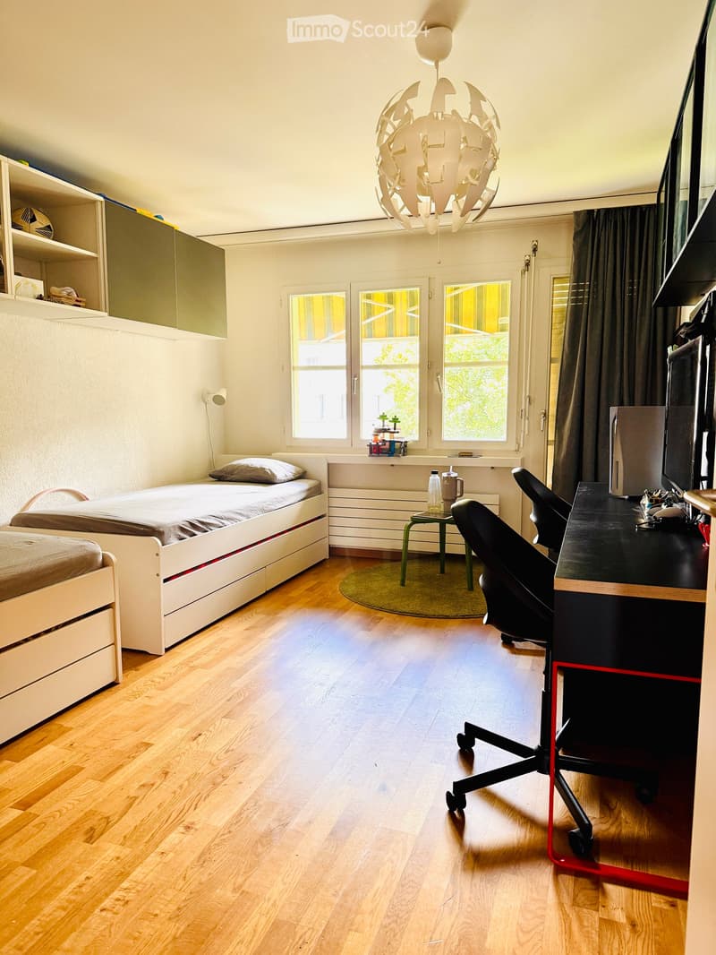 Modern bedroom with wooden flooring, a single bed, desk, chair, small round stool, built-in cabinet, shelves, and a decorative hanging lamp.