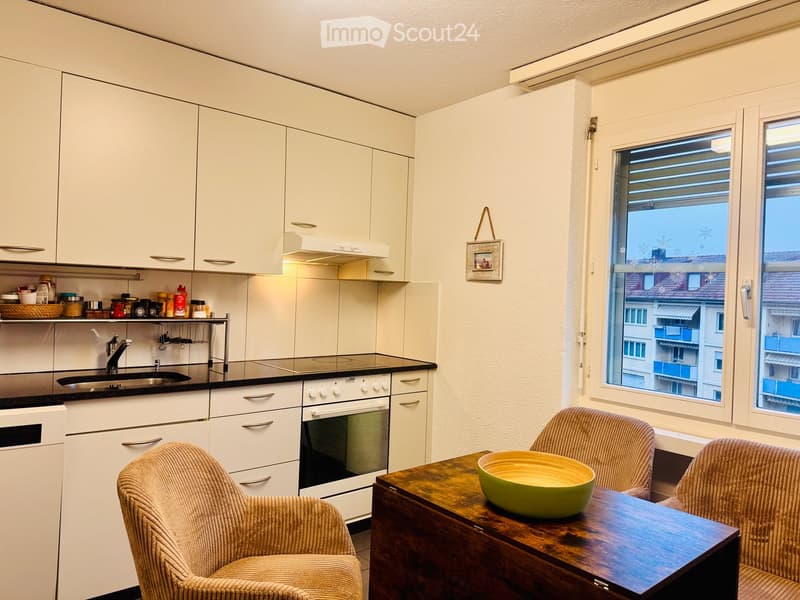 A modern kitchen with white cabinets, a wooden dining table, two chairs, and a green bowl on the table. The kitchen features a sink, oven, and various small appliances on the counter.