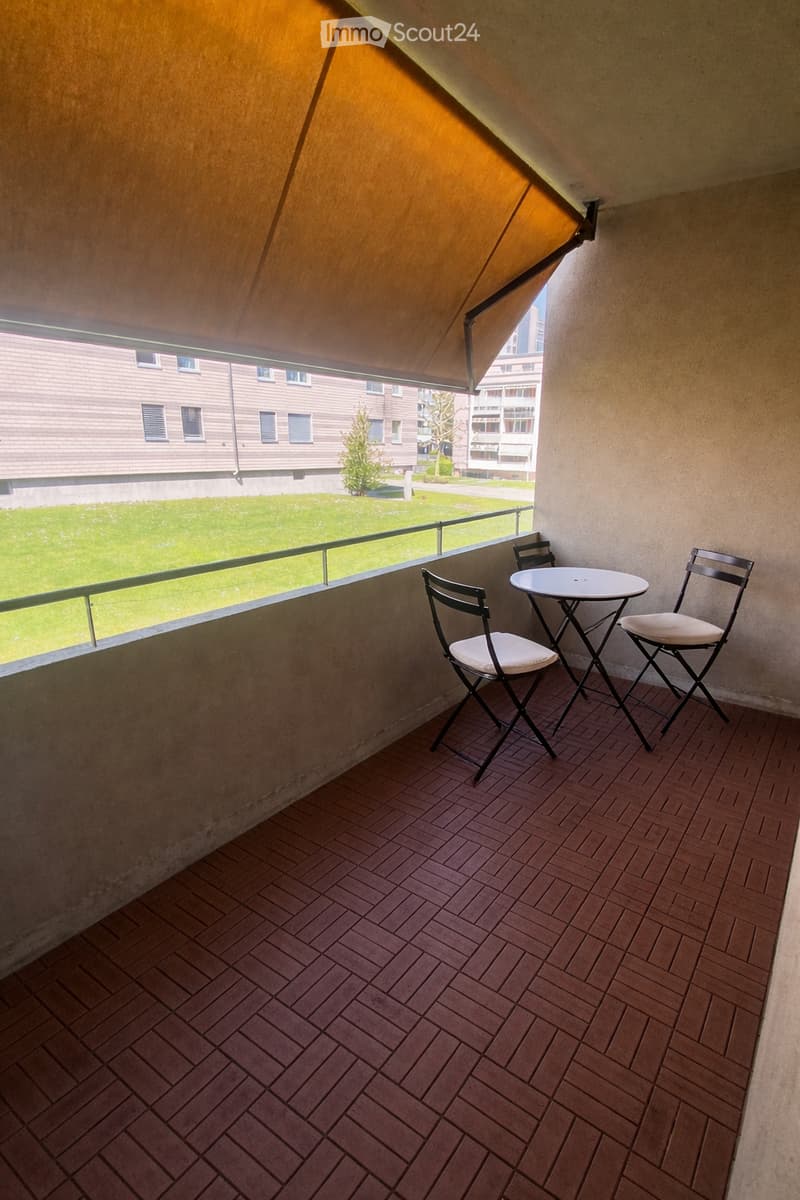 Balcony with wooden decking, small round table, and two folding chairs, overlooking a green lawn and neighboring buildings.