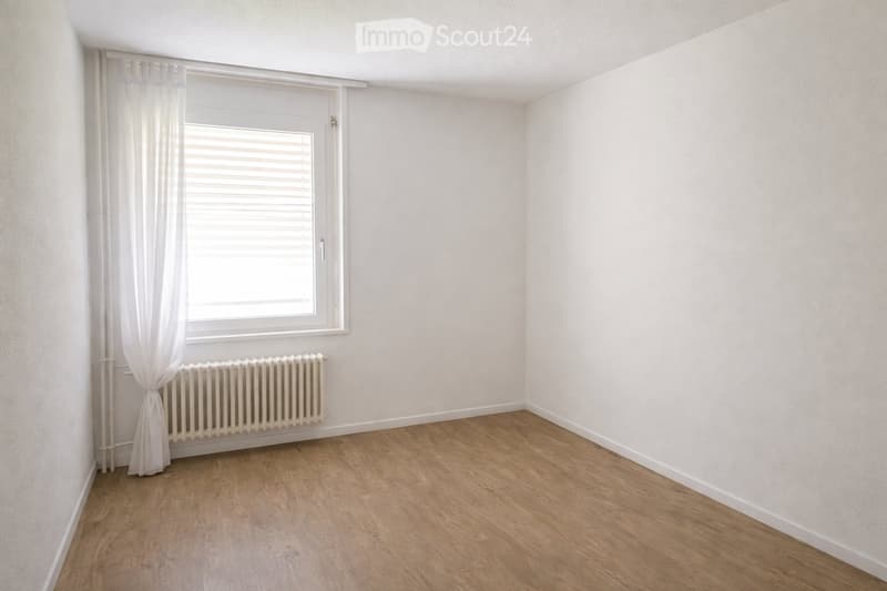 Empty room with wooden flooring, white walls, a window with white curtains, and a radiator below the window.